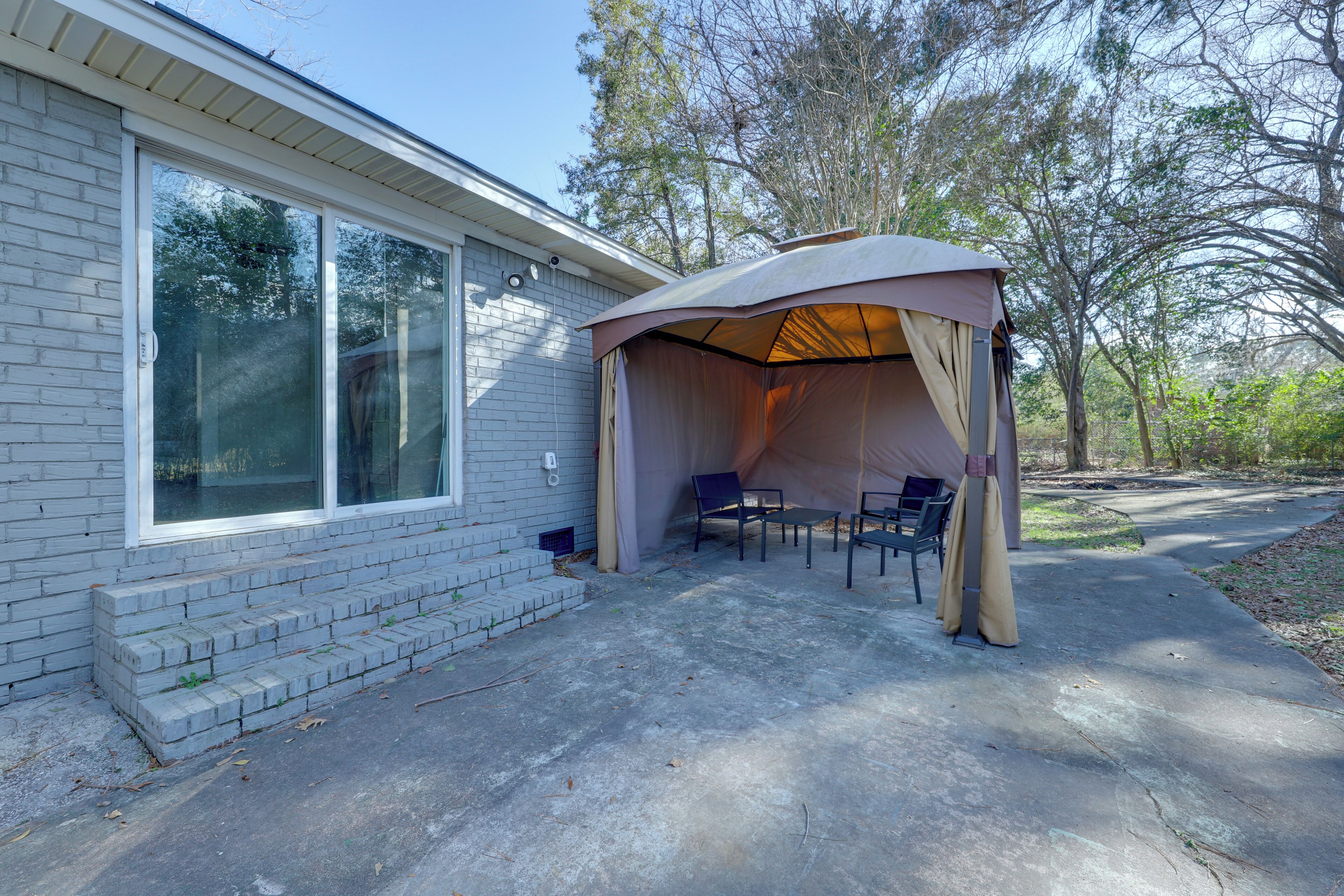 A covered patio area with a table and chairs, a brick house, and trees in the background.