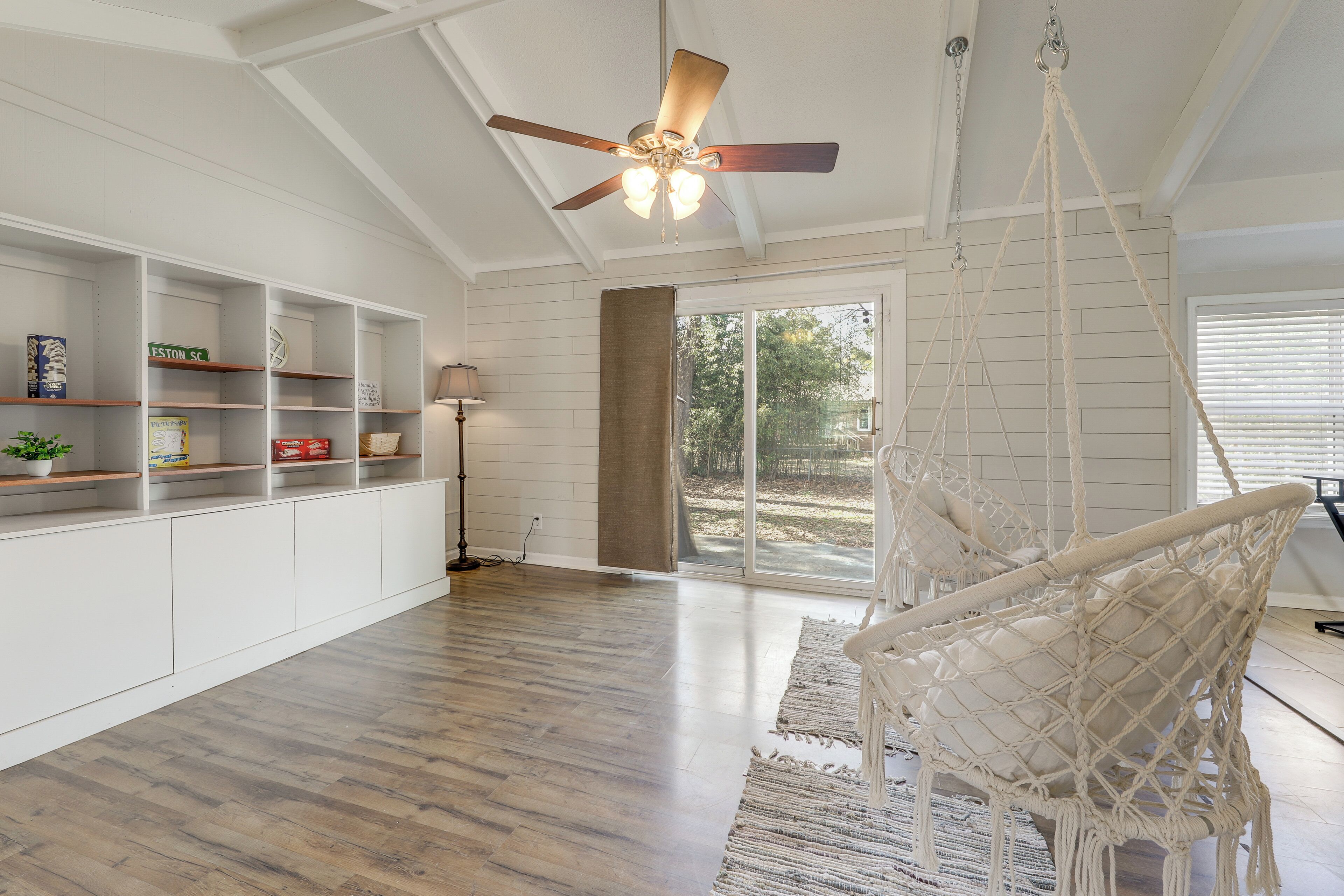 Una habitación amplia con piso de madera, paredes blancas, un ventilador de techo, un ventanal grande y una silla de macramé.