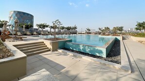 Un área de piscina moderna con un edificio geométrico al fondo, rodeada de sillas de descanso y escalones que dan a la piscina.