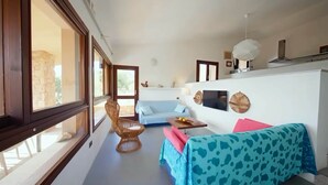 Een knusse woonkamer met een blauwe bank, een rieten fauteuil, een tafeltje met fruit, en een aan de muur bevestigde televisie.