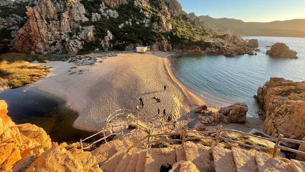 Een kustlandschap met een rotsachtig strand, een houten trap die naar het water leidt en een klein gebouw in de verte.