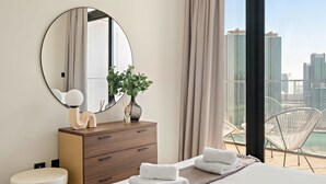 Een moderne hotelkamer met een grote ronde spiegel, een houten dressoir, een rond witte kruk, een bed met keurig opgevouwen witte handdoeken, en een balkon met uitzicht op de skyline van de stad en een wateroppervlak.