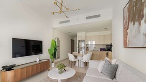 A modern living room with a flat-screen TV, a wooden console table, a round coffee table with a plant, a grey sofa with cushions, and a dining area with a white table and chairs. - Silkhaus Luxurious 1BR Downtown Burj Royale (Dubai)