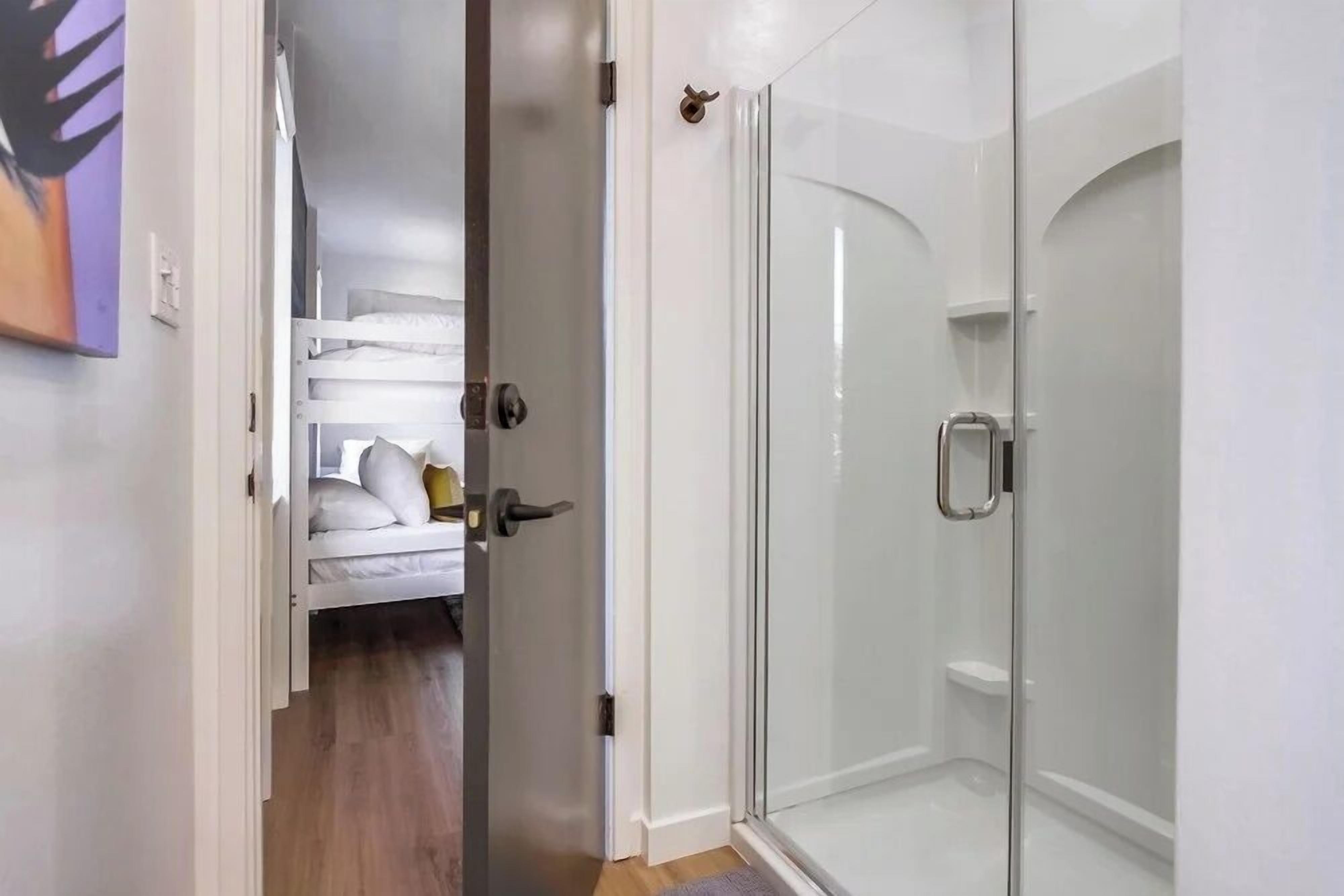 A modern bathroom with a glass shower enclosure and a white door leading to a bedroom with a bunk bed.