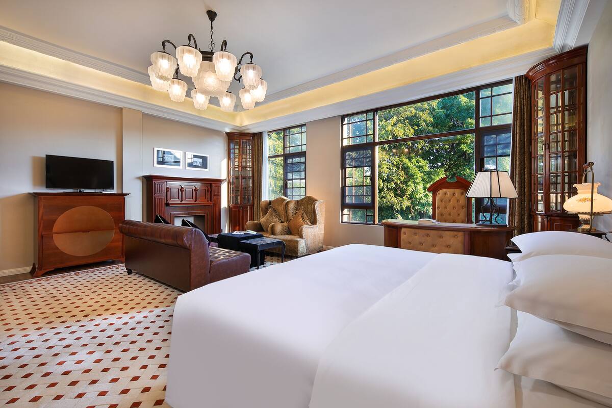 A hotel room with a large bed, a sofa, a television, a cabinet, and a window with a view of trees.