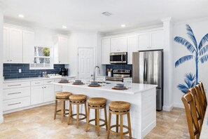 A modern kitchen with white cabinetry, a large island with stools, stainless steel appliances, and a blue palm tree mural.