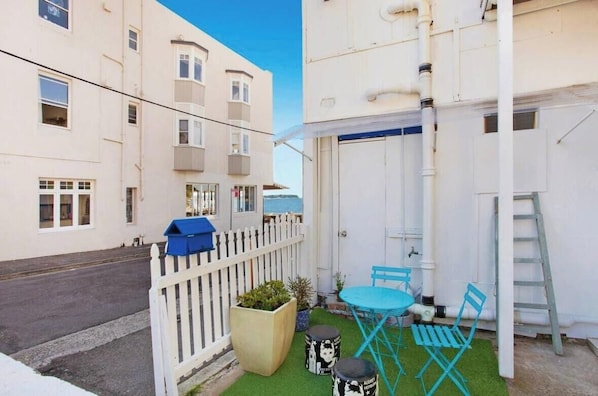 Eine kleine Terrasse im Freien mit Tisch und Stühlen, einem weißen Zierzaun und einem Gebäude mit mehreren Fenstern und Balkonen.