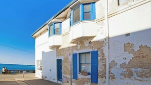 Un bâtiment situé sur la côte, avec une peinture blanche écaillée et des volets bleus, une porte bien visible et une petite fenêtre ornée d’une plante.