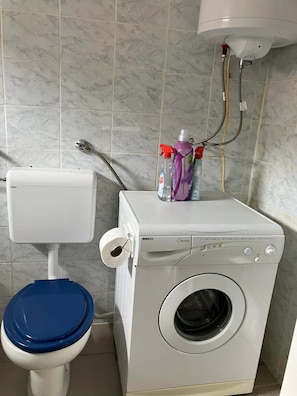 A compact bathroom with a toilet, a washing machine, and a shower unit. - 2BR Apartment With Garden in Povljana (Povljana)
