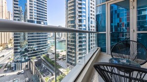 A balcony with a glass railing overlooking a cityscape with high-rise buildings and a river. There are two black wicker chairs and a small glass table on the balcony. - Silkhaus Dubai Marina Studio Bay Central Tower (Dubai)