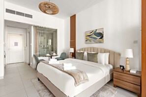 Een moderne hotelkamer met een groot bed, een douchecabine van glas, een zithoek met een stoel en een nachtkastje van hout met een lamp.