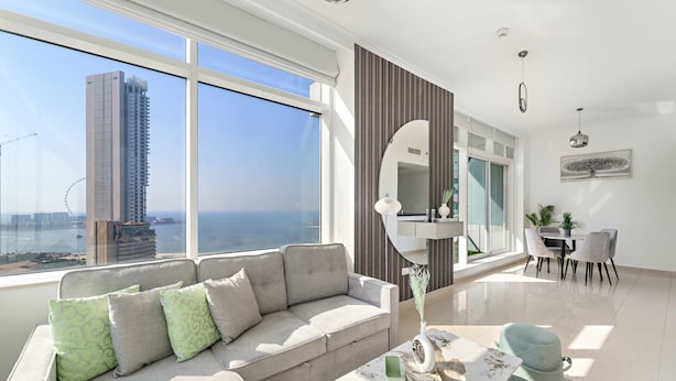 Una sala de estar moderna con un ventanal grande, un sofá, un comedor y vistas a un edificio alto y al mar.