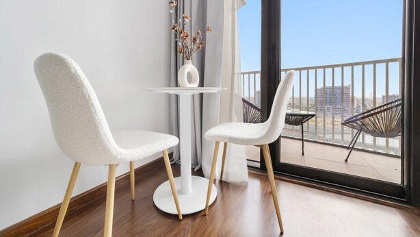 A modern room with two white chairs, a small white table, and a vase with dried flowers. A balcony with outdoor furniture is visible through a large window. - Silkhaus Cozy Studio Balcony Binghatti Pearl (Dubai)