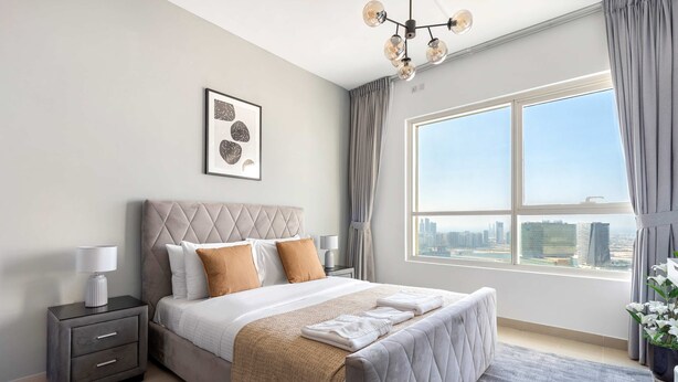 A modern bedroom with a large bed, a nightstand, a framed artwork, and a view of the city through a large window. - Silkhaus Cozy 1BR With Balcony Pool Gym on Reem Island (Abu Dhabi)