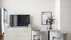 A modern living room with a large flat-screen TV, a white console with geometric patterns, two grey armchairs, a round table, and a framed abstract painting. - Silkhaus New High Floor Studio Aykon City (Dubai)