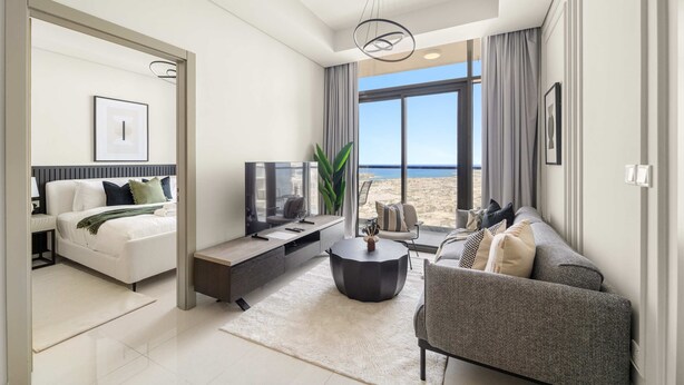 A modern living room with a sofa, coffee table, and TV, a large window with a beach view, and a bed with a nightstand. - Silkhaus Elite 2bdr Near Safa Park Aykon City (Dubai)