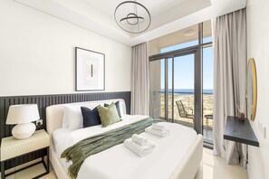 Een moderne hotelkamer met een groot bed, een nachtkastje met lamp, uitzicht op het strand en een balkon met tafel en stoelen.