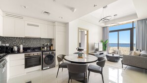 A modern kitchen with white cabinets, a black countertop, and a built-in oven. There is a dining area with a round table and grey chairs. A large window offers a view of the beach. - Silkhaus Elite 2bdr Near Safa Park Aykon City (Dubai)
