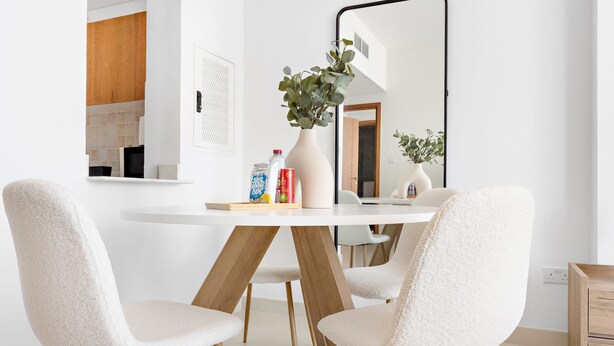 Un espace repas moderne avec une table blanche, quatre fauteuils moelleux, un vase avec des plantes vertes et un grand miroir au mur.