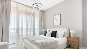 Une chambre moderne avec un grand lit, une table de chevet avec une lampe et une vue sur l’extérieur à travers des rideaux légers.