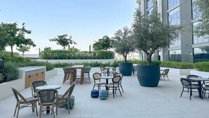 An outdoor patio area with tables and chairs, surrounded by greenery and modern buildings. - Silkhaus Cozy 2 BR Full Amenities (Dubai)