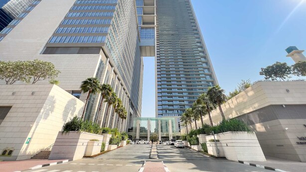Un immeuble moderne de grande hauteur, doté d’une façade en verre remarquable, entouré de palmiers et bordé d’une promenade dallée.