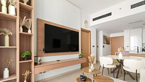 Ett modernt vardagsrum med en stor tv-skärm, trähyllor med dekorativa föremål, en matplats med ett vitt bord och stolar samt ett köksområde i bakgrunden.