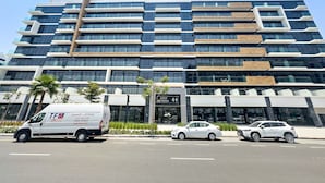 A modern multi-story building with a mix of glass and wood facade, a prominent entrance with a sign, and a parking area with vehicles. - Silkhaus Chic Studio Skyline View MBR City (Dubai)