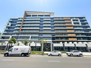 Ein modernes, mehrgeschossiges Gebäude mit einer Fassade aus Glas und Holz, einem markanten Eingang mit Beschilderung und einem Parkplatz mit Fahrzeugen.
