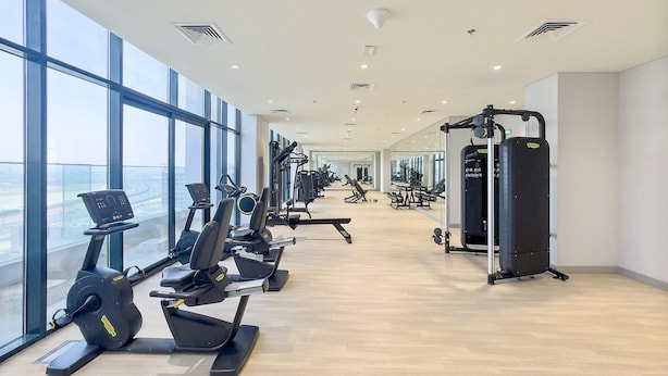 A modern gym with cardio machines, weightlifting equipment, and large windows offering views of the city. - Silkhaus Chic Studio Skyline View MBR City (Dubai)
