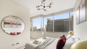 Quarto de hotel moderno com uma cama grande, um espelho redondo e vista para a cidade através de grandes janelas.