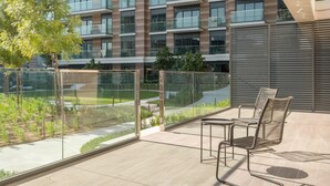 Een modern zitgedeelte buiten met uitzicht op een meerhoog gebouw, een glazen balustrade en een boom.