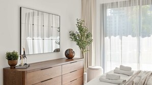 Een moderne slaapkamer met een houten dressoir, een grote spiegel, een potplant en een netjes opgemaakt bed met handdoeken.
