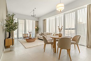 Uma sala de estar moderna com sofá, mesa de jantar, cadeiras, uma grande planta em vaso e janelas amplas com cortinas leves.