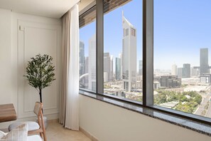 A room with a large window offering a cityscape view, a potted plant, and a seating area with a wooden chair and a cushioned seat. - Silkhaus Urban 1bdr Heart of Difc Sky Gardens (Dubai)