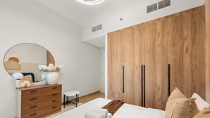 Un dormitorio moderno con un armario de madera, un espejo redondo, una mesita de noche con un jarrón y una cama bien hecha.
