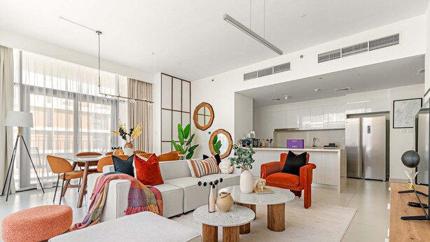 A modern living room with a sofa, ottomans, coffee table, and a kitchen in the background. - Silkhaus Luxurious 3BR Maid Dubai Hills (Dubai)