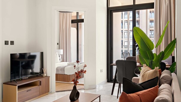 A modern living room with a flat-screen TV on a wooden stand, a coffee table with a vase, a sofa with cushions, and a large window with a view of buildings. - Silkhaus 1bdr Prime Location in Maryah Island (Abu Dhabi)