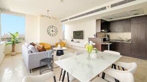 Ein modernes Wohnzimmer mit Sofa, Esstisch und Stühlen. An der Wand ist ein Fernseher befestigt, und es gibt eine Küchenzeile mit Schränken und Geräten.