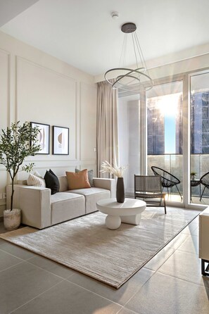 A modern living room with a sofa, coffee table, and chairs, large windows with a city view, and a hanging light fixture. - Silkhaus Modern 1 BDR Forte Tower Downtown (Dubai)
