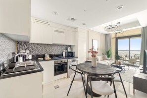 A modern kitchen with white cabinets, a black countertop, and a dining area with a round table and chairs. - Silkhaus Stylish 2bdr in Aykon Tower (Dubai)