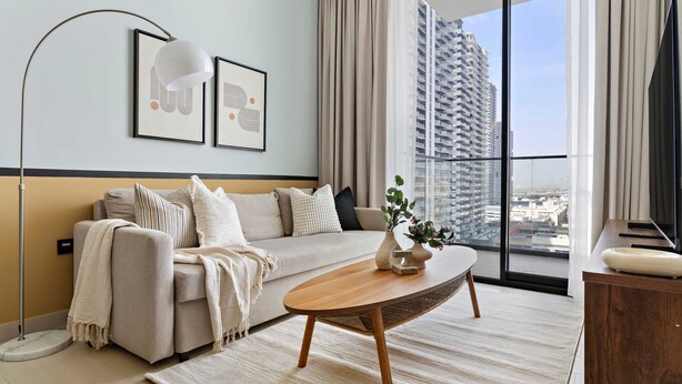 A modern living room with a sofa, a coffee table, a floor lamp, and a large window with a city view. - Silkhaus Elite 1bdr in JVC Summer Promotion (Dubai)