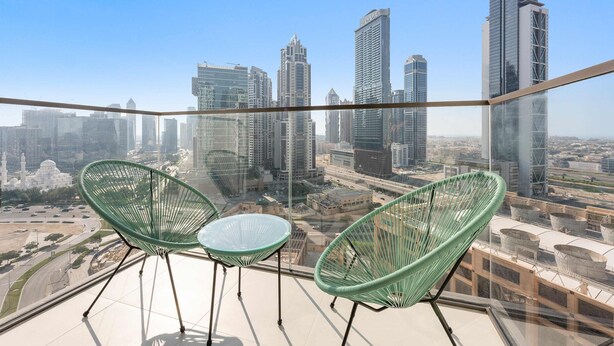 Ein Balkon mit zwei grünen Stühlen und einem kleinen Tisch, mit Blick auf eine Skyline und moderne Wolkenkratzer.