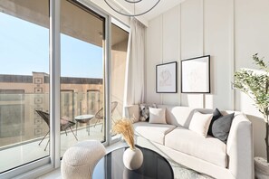A modern living room with a sofa, a coffee table, and a vase with dried plants. There are two framed artworks on the wall and a balcony with outdoor furniture. - Downtown Apartment w Tower Pool Balcony (Dubai)