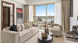 Uma sala de estar moderna com um sofá, poltronas e uma mesa de centro. Há uma janela grande com vista para um horizonte urbano e para a água.