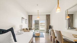 A modern hotel room with a bed, a sitting area with a sofa and chairs, a dining table, and a large window with a view of buildings. - Silkhaus Studio Masdar City Leonardo Residence (Abu Dhabi)