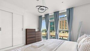 A modern hotel room with a large bed, a wooden dresser, and a view of a cityscape through large windows. - Silkhaus Chic 1bdr in Al Bandar Al Naseem A (Abu Dhabi)