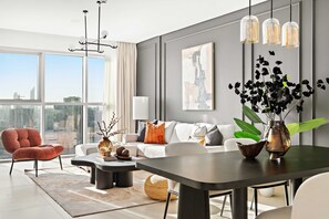 A modern living room with a sofa, armchair, coffee table, and dining table. There is a large window with a view of the city. - Silkhaus Canal View 1BR Apartment In Reem Island (Abu Dhabi)