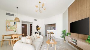 Una sala de estar moderna con sofá, una mesa de centro y un área de comedor. Hay un televisor montado en la pared y una cocina al fondo.