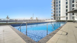A swimming pool with a tiled deck, surrounded by a white fence, and a tall building in the background. - Silkhaus Large 1bdr Near Yas Island Parks (Abu Dhabi)